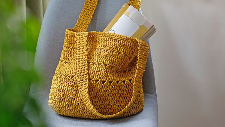Gelbe gehäkelte Tasche mit zwei Henkeln liegt auf einem grauen Stuhl, gefüllt mit Büchern und Papieren.