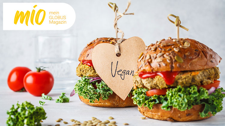 Zwei vegane Hamburger mit Salat, Tomaten und Ketchup, ein Herz-Anhänger mit „Vegan“, daneben ein Glas Wasser und zwei Tomaten.