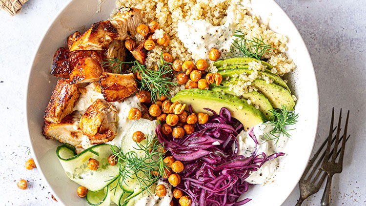 Weiße Schüssel mit Quinoa, gegrilltem Hähnchen, Avocado, Gurke, roten Zwiebeln, gerösteten Kichererbsen und Dill.