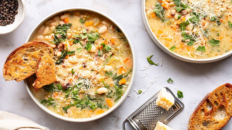 Zwei Schalen mit Gemüsesuppe, dekoriert mit Kräutern und geriebenem Käse, daneben geröstetes Brot und eine Käsereibe.