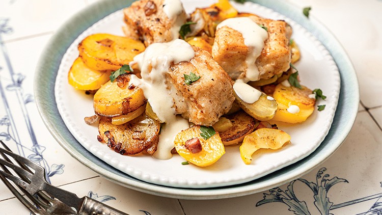 Ein Teller mit gebratenen Kartoffelscheiben, panierten Fischwürfeln, heller Sauce und dekorativer Petersilie.