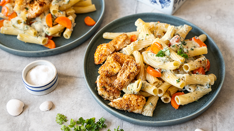 Zwei blaue Teller mit Rigatoni, heller Soße, Karottenscheiben und panierten Hähnchenstücken auf hellgrauem Tisch.