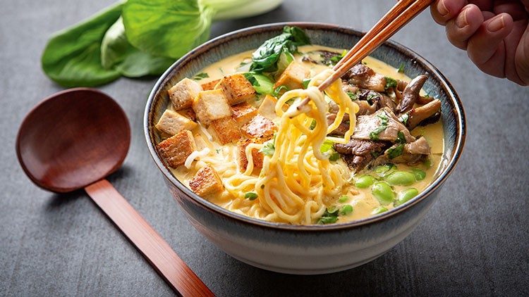 Schüssel mit gelben Nudeln in heller Brühe, Tofu-Würfeln, Blattgemüse, Pilzen und Edamame, daneben ein Holzlöffel auf grauem Untergrund.