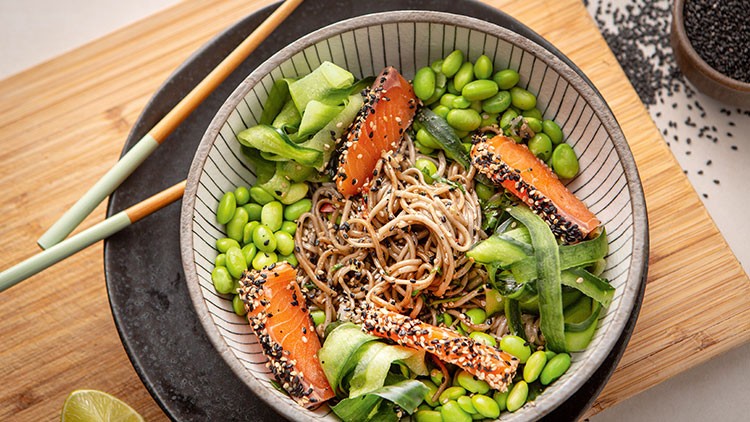 Bunte Bowl mit Soba-Nudeln, Edamame, Gurkenstreifen und Sesam-Lachs auf einem schwarzen Teller mit Essstäbchen.