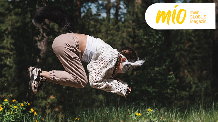Person in beiger Hose und gemustertem Pullover springt im Wald, rechts oben gelbes mio-Logo mit Text „mein GLOBUS Magazin“.