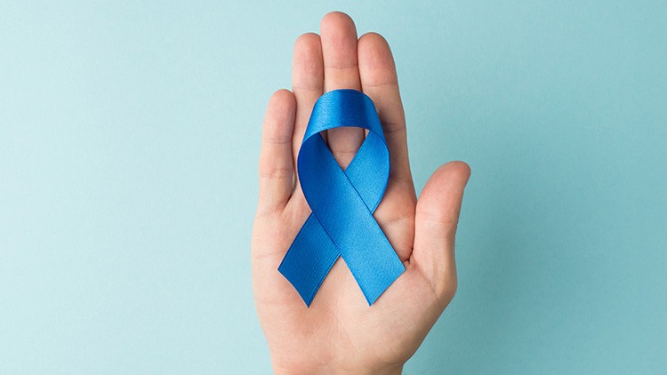 Eine Hand hält eine blaue Schleife vor einem hellblauen Hintergrund.