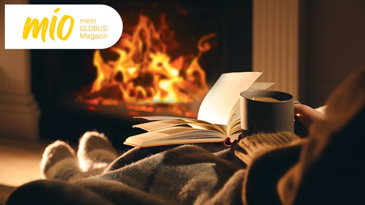 Geöffneter Buchdeckel, graue Tasse, warmes Feuer im Hintergrund und Schriftzug mio mein GLOBUS Magazin links oben.
