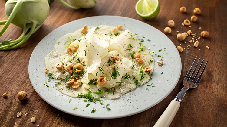 Auf einem Teller liegt dünn geschnittenes Kohlrabi-Carpaccio, garniert mit Kräutern, Haselnüssen und gehobeltem Parmesan.
