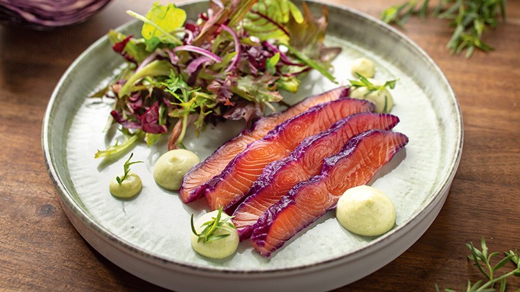 Dünne Scheiben gebeizter Lachs, bunter Blattsalat und vier grüne Kräutercremetupfer auf einem hellen Teller.