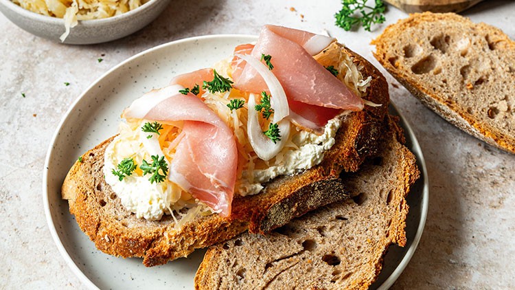 Rustikale Brotscheiben mit Frischkäse, Sauerkraut, Schinken und Petersilie auf einem weißen Teller, daneben weitere Brotscheiben.