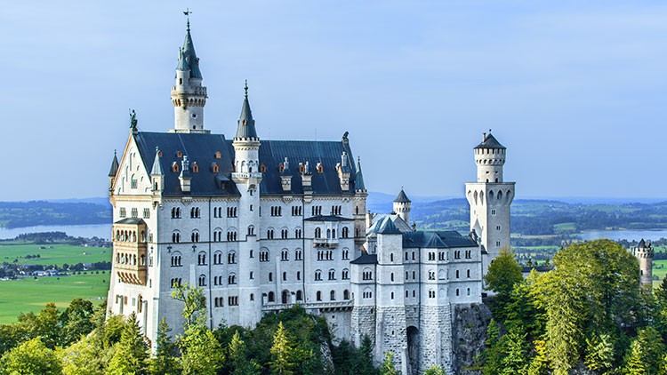 Schloss Neuschwanstein mit weißen Fassaden und mehreren Türmen vor einer grünen Landschaft und blauem Himmel.