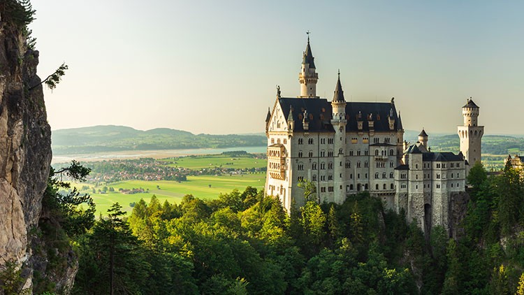 Schloss Neuschwanstein mit spitzen Türmen liegt erhöht am Waldrand, umgeben von grünen Wiesen und einem weiten Tal im Hintergrund.