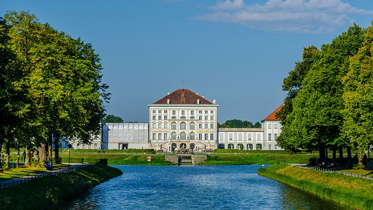 Helles Schloss mit symmetrischem Wasserkanal, flankiert von grünen Baumreihen unter blauem Himmel mit wenigen Wolken.