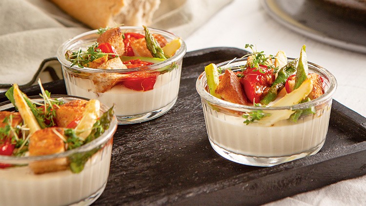 Drei Glasförmchen mit weißer Creme, garniert mit grünem Spargel, Brotwürfeln, roten Tomaten und frischen Kräutern auf schwarzem Tablett.