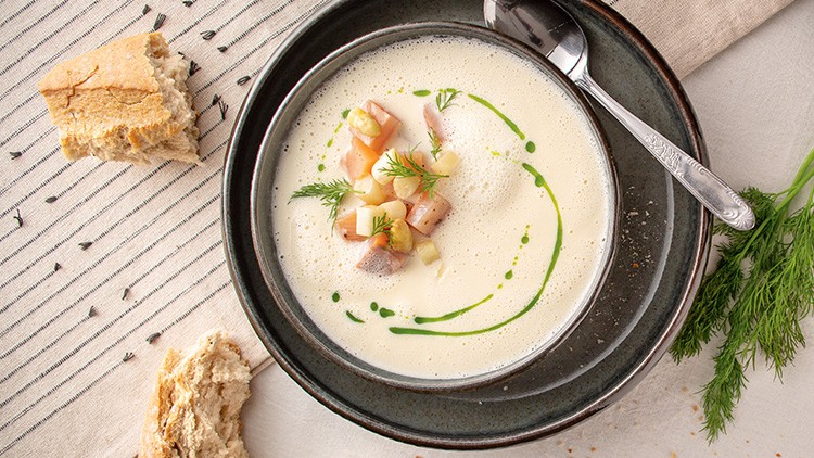 Cremige Suppe mit Kräuterdekoration, Gemüse- und Fischstücken in einer dunklen Schale, daneben Brot und ein Löffel.