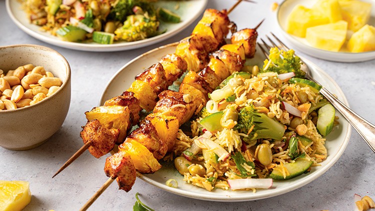 Zwei gegrillte Spieße mit Hähnchen und Ananas neben einem Teller mit Reis, Gemüse und Nüssen auf einem hellen Tisch.
