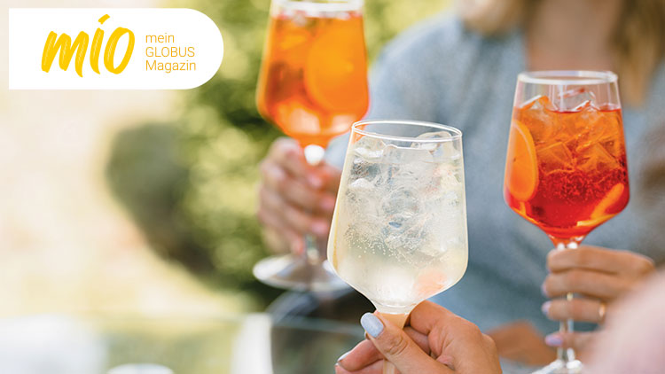 Drei Gläser mit verschiedenen Getränken, darunter Eistee und Aperol, vor einer unscharfen grünen Naturkulisse, Schriftzug: mio mein GLOBUS Magazin.