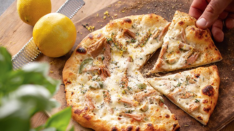 Frisch gebackene Pizza mit Hähnchenstücken, frischen Kräutern und Käse, daneben zwei Zitronen und eine Reibe auf Holzuntergrund.