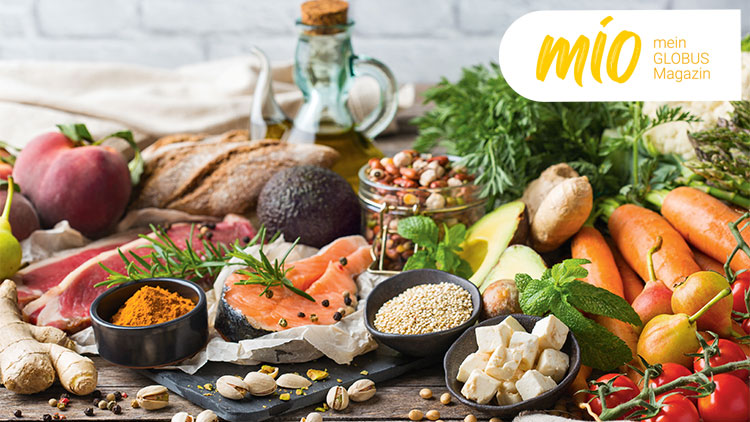 Frische Lebensmittel wie Lachs, Gemüse, Käse, Gewürze und Nüsse auf einem Tisch, daneben „mio mein GLOBUS Magazin“.
