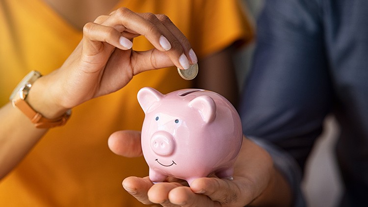Eine Hand hält ein rosa Sparschwein, während eine andere Hand eine Münze in den Schlitz des Sparschweins steckt.