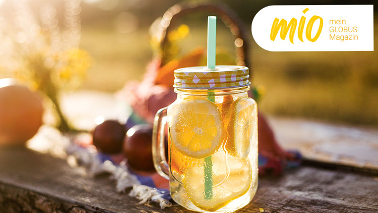 Ein Glas mit gelber Flüssigkeit, Zitronenscheiben und grünem Strohhalm steht auf einem Holztisch, daneben steht „mio mein GLOBUS Magazin“.