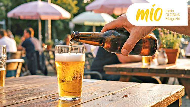 Eine Hand schenkt Bier aus einer Flasche in ein Glas ein, im Hintergrund sind Menschen und Sonnenschirme erkennbar, Text: mio mein GLOBUS Magazin.