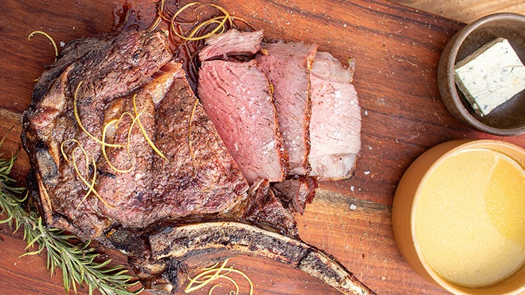 Gegrilltes Rinderkotelett mit frischen Kräutern, geschnittenem Fleisch, einem hellen Dip und einer Kräuterbutter auf Holzbrett.