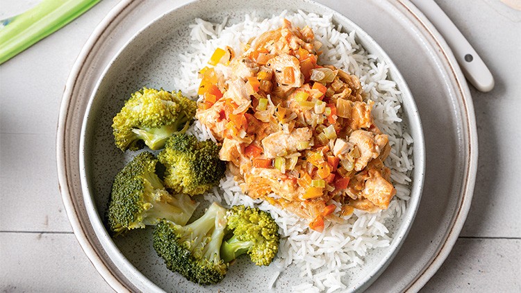 Gekochter Reis mit Hähnchen-Gemüse-Mischung in heller Sauce und Brokkoli auf einem grauen Teller, daneben ein weißes Besteck.