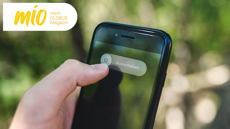Finger zeigt auf Smartphone-Bildschirm mit Button und Text „Ausschalten“, links oben steht „mio mein GLOBUS Magazin“.