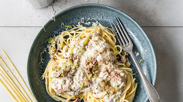 Ein Teller mit Spaghetti, heller Soße, Thunfisch und Lauch, daneben ein Besteck und rohe Spaghetti auf grauer Oberfläche.