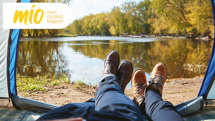 Zwei Personen sitzen mit Wanderstiefeln vor einem Zelt an einem See, oben steht „mio mein GLOBUS Magazin“ in gelber Schrift.
