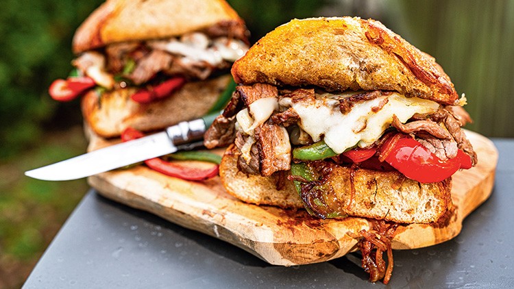 Frisches Sandwich mit gegrilltem Fleisch, geschmolzenem Käse, Paprika und Zwiebeln auf einem Holzteller mit Messer.