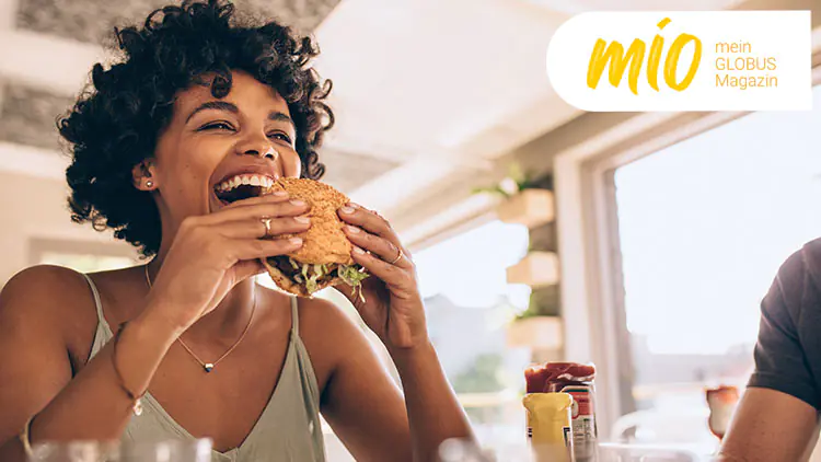 Eine Person hält einen Burger mit Sesambrötchen und Salat, oben rechts steht „mio mein GLOBUS Magazin“ in Gelb und Weiß.