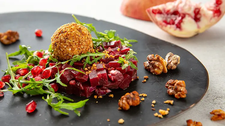 Rote Bete-Würfel mit Rauke, Granatapfelkernen, Walnüssen und einem panierten Ball auf einem schwarzen Teller.