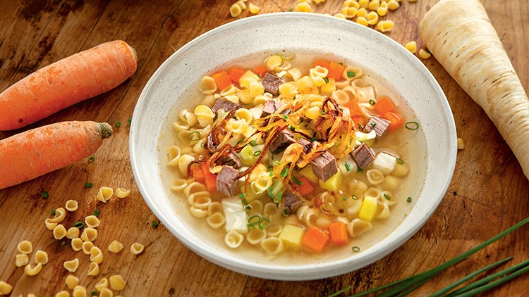 Ein weißer Teller mit Suppe, gefüllt mit Nudeln, Fleischwürfeln, Karotten, Petersilienwurzel, Lauch und Röstzwiebeln, dekoriert mit Schnittlauch.