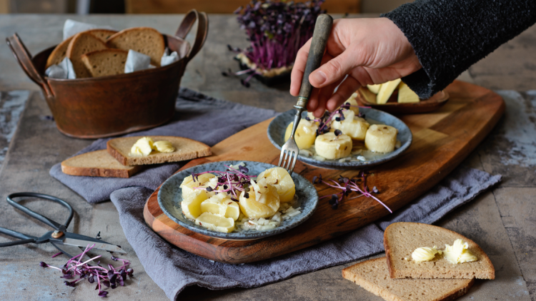 Drei Teller mit Kartoffelscheiben, Zwiebeln und Kräutern, daneben Brot mit Butter, eine Hand hält eine Gabel.