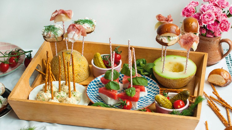 Eine Holzbox mit Snacks auf Spießen, darunter Melone mit Käse, Brot, Schinken, Minipretzeln, Gurken und Tomaten.