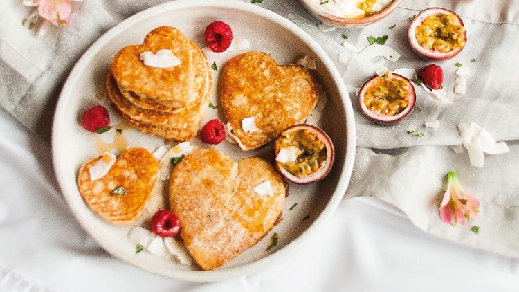 Herzförmige Pancakes mit Himbeeren und Passionsfrüchten neben Smoothie, Kaffee und Joghurt auf hellem Tisch mit Blumen.
