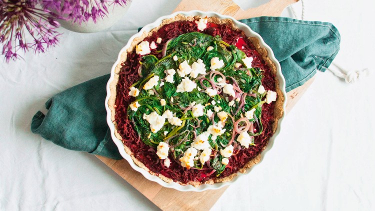 Eine Quiche mit grünem Blattgemüse, roten Zwiebeln und weißem Käse auf einem Holzbrett mit grünem Stofftuch.