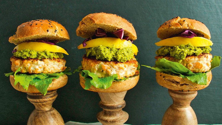Drei Mini-Burger mit Brötchen, Salat, Patties, gelbem Belag, grünem Aufstrich und Rotkohl auf hölzernen Sockeln vor dunklem Hintergrund.