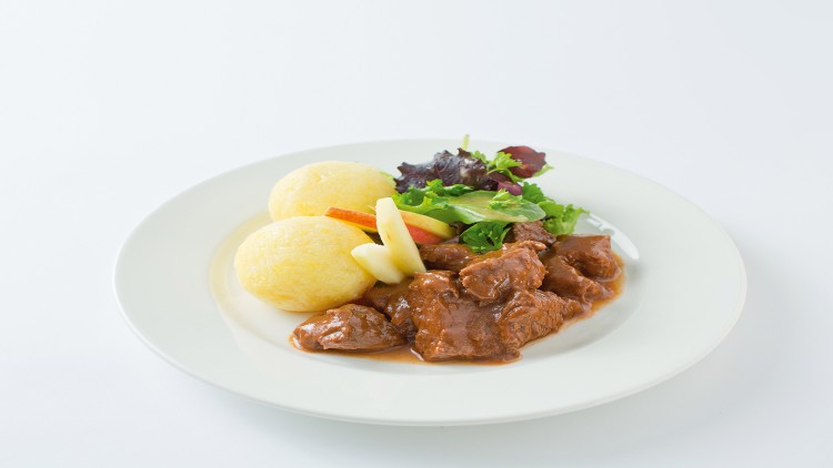 Teller mit zwei Kartoffelklößen, Fleisch in brauner Sauce, gemischtem Salat und Apfelspalten auf weißem Untergrund.