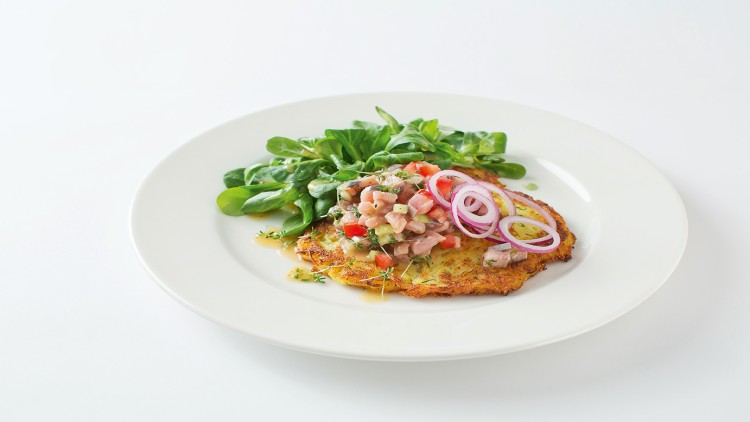 Ein Rösti auf weißem Teller, belegt mit Tatar, roten Zwiebeln und Feldsalat, garniert mit frischen Kräutern.