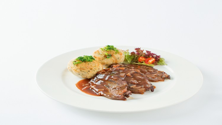 Zwei Scheiben Braten mit dunkler Soße, zwei Semmelknödeln mit Petersilie und Salatgarnitur auf einem weißen Teller.