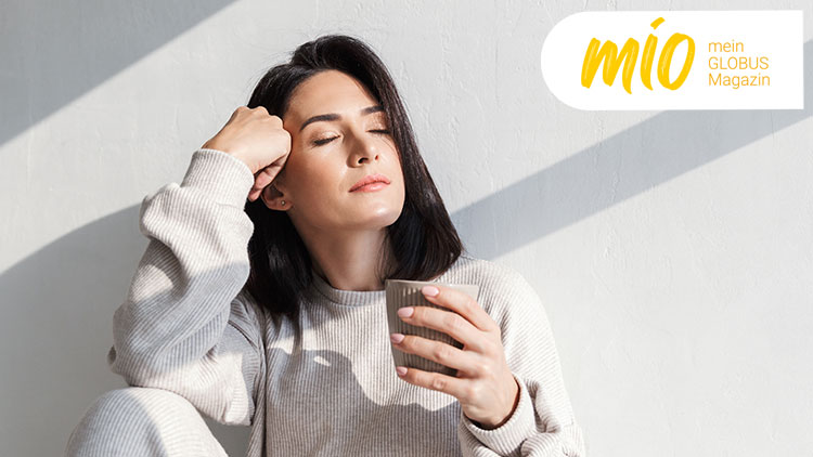Person in beigem Pullover hält eine Tasse vor heller Wand, oben rechts das mio mein GLOBUS Magazin-Logo in Gelb.