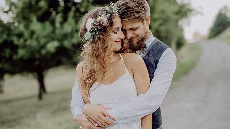 Person in weißem Kleid mit Blumen im Haar wird von einer Person in Hemd und Weste auf einem Feldweg umarmt.