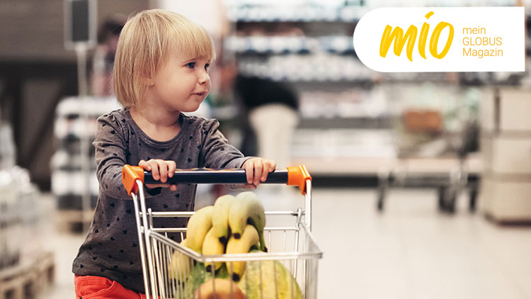 Ein Kind schiebt einen Einkaufswagen mit Bananen durch einen Supermarkt, oben rechts steht „mio mein GLOBUS Magazin“.