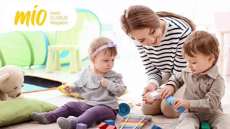 Drei Personen spielen mit bunten Spielsachen auf einem weichen Teppich, daneben ein weißer Teddybär, im Hintergrund steht mio mein GLOBUS Magazin.