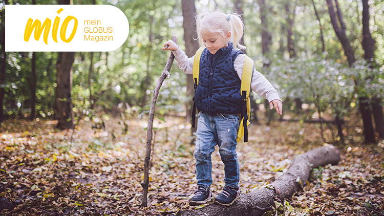 Kind im Wald mit gelber Weste und Stock in der Hand, links oben das mio mein GLOBUS Magazin Logo auf weißem Hintergrund.