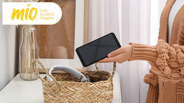 Eine Hand in beigem Strickpullover legt ein schwarzes Smartphone in einen geflochtenen Korb mit weißen Kopfhörern, mio mein GLOBUS Magazin.