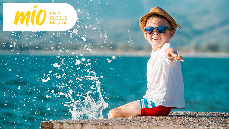 Kind in weißem Oberteil und Strohhut am Wasser, spritzendes Wasser, Schriftzug „mio mein GLOBUS Magazin“ oben links.